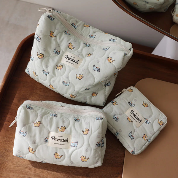 A white quilted cosmetic storage pouch with a light blue and yellow cat print is shown on a wooden surface.