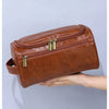 A brown leather toiletry bag with a zipper closure is held in a person's hand.