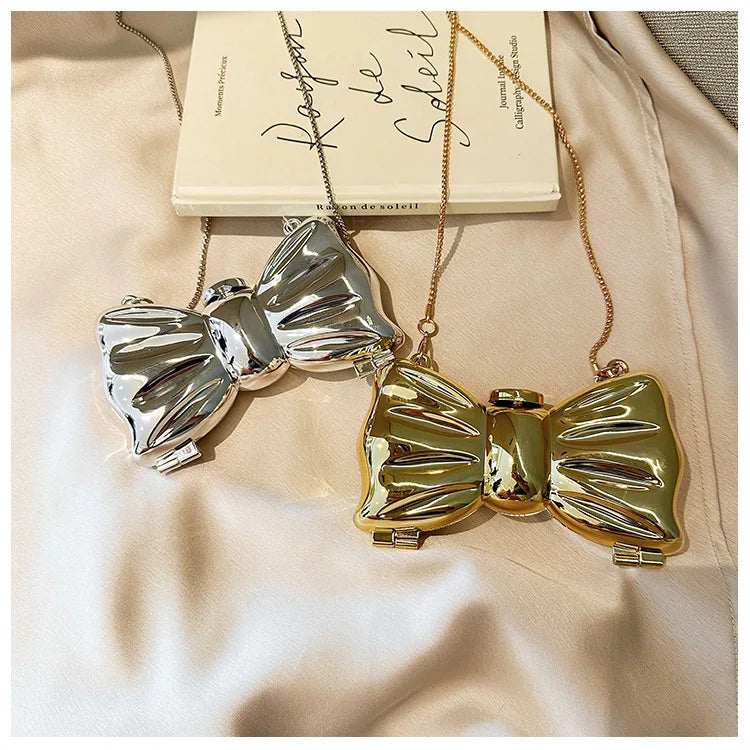 A gold-toned bow knot evening bag with a silver bow detail is displayed against a light beige fabric next to a book.