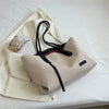 A beige canvas shoulder tote bag with black straps and a small clear charm resting on a white cloth.