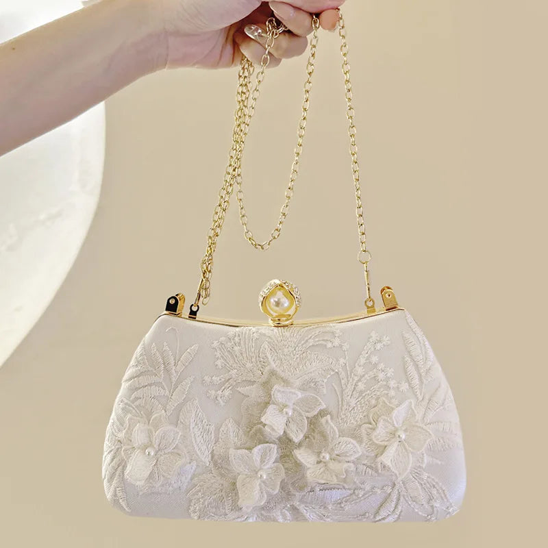 A white clutch bag with delicate pearl embroidery and a gold chain strap being held by a hand.