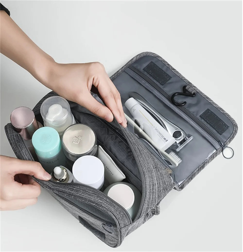 A hand is opening a gray portable waterproof toiletry washbag containing various personal care items.