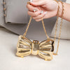 A close-up of a gold acrylic bow clasp mini handbag with a chain strap being held by a hand.