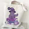 A canvas tote bag featuring a purple cat illustration sitting atop a stack of books and a teacup.