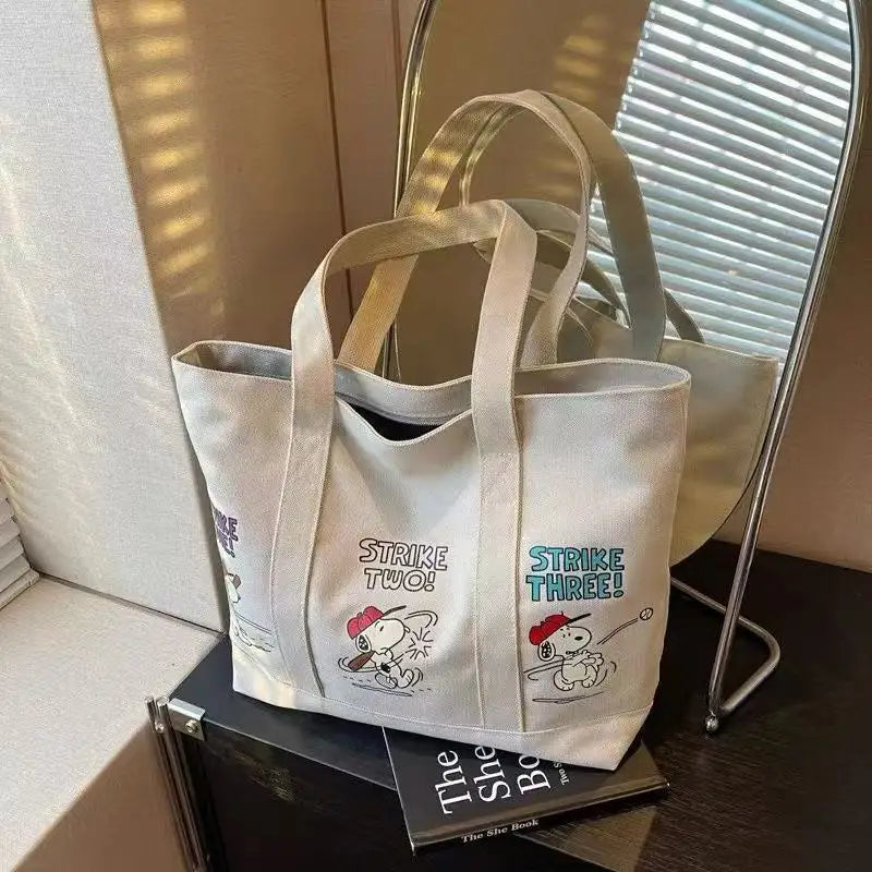 A white canvas shoulder bag featuring a Snoopy cartoon design with the text Strike Two Strike Three