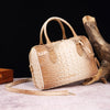 A light tan crocodile pattern leather crossbody tote bag resting on a pink surface next to a piece of driftwood.
