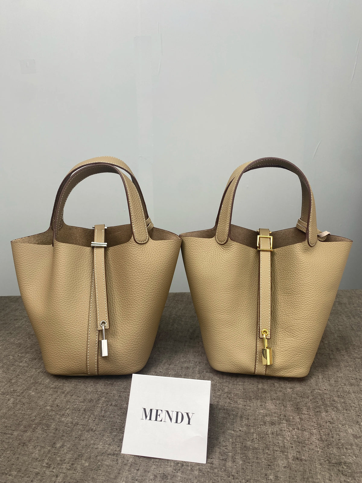 A beige genuine leather bucket bag with a drawstring closure and gold hardware displayed on a gray surface.