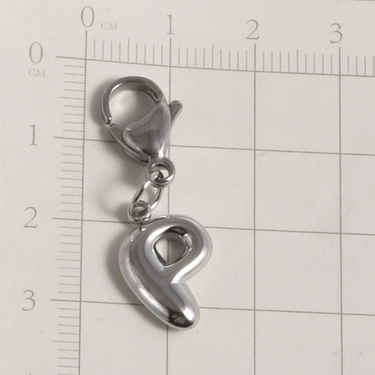 Silver stainless steel alphabet letter charm with a lobster clasp placed on a ruler.