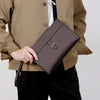 A hand holds a dark brown men's PU leather clutch bag against a beige jacket and black pants.