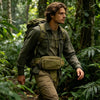 A man wearing a tactical concealed carry waist pack and olive green shirt walks through a dense, lush forest.