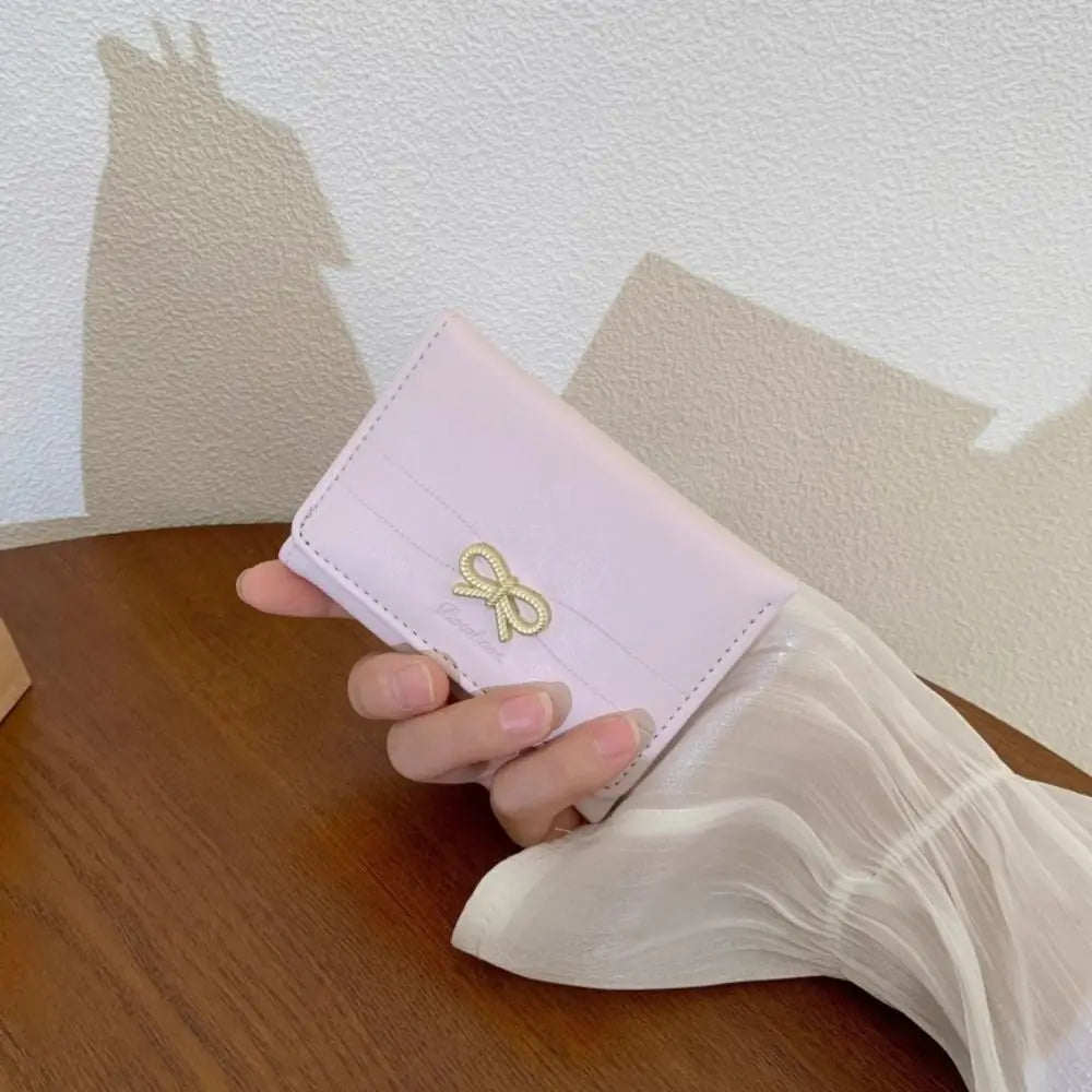 Une main tient un porte-cartes tri-pli rose pâle en PU avec un charm en torsade dorée sur une surface en bois.