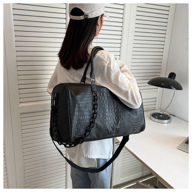 A person is holding a black crocodile grain PU leather travel duffle bag in front of white slatted blinds.