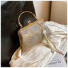 A light gray embroidered silk evening clutch with a gold chain strap resting on white fabric.