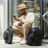 A man wearing a beige hat sits next to a black backpack and a smartphone in a modern setting.