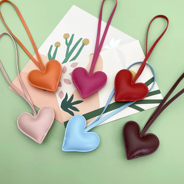 A collection of five heart-shaped PU leather keychains in various shades of pink, red, purple, blue, and brown on a light gre