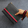 Schwarze, gemusterte Clutchtasche mit roten und grünen Streifen wird von einer Hand auf einer grauen Stoffoberfläche gehalten