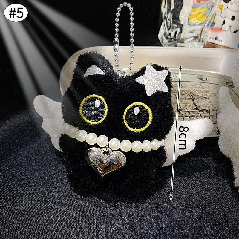 Colgante de peluche de gato negro con ojos dorados y un dije de corazón plateado, exhibido en una bandeja blanca.