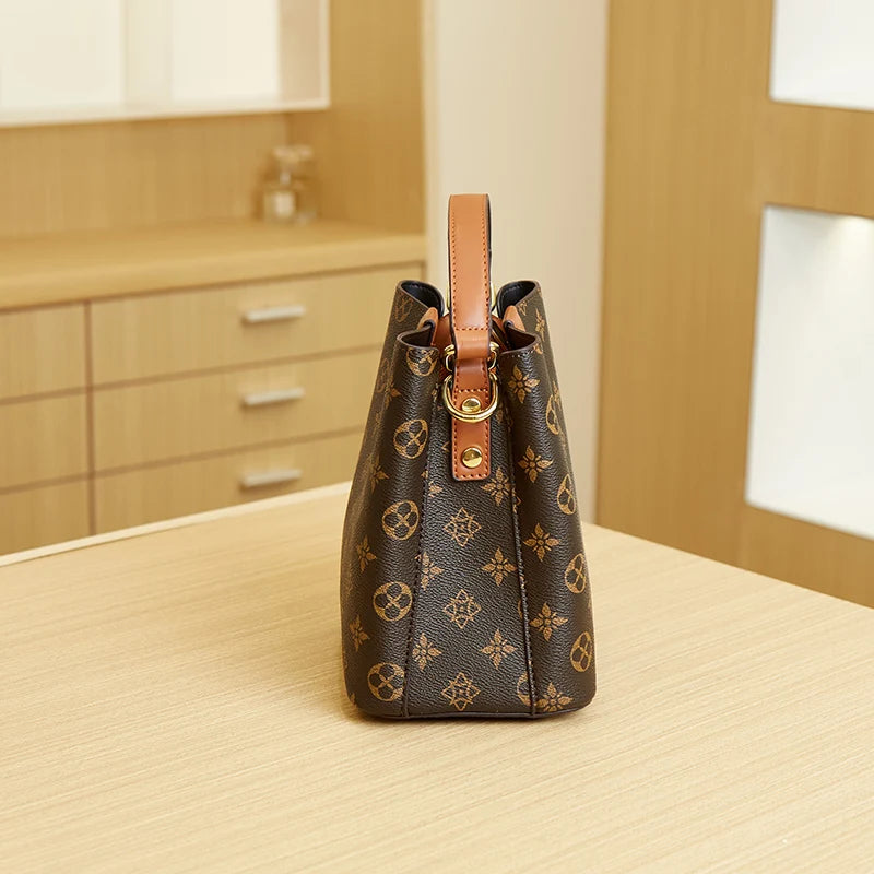A brown leather bucket bag with a floral pattern and a tan adjustable strap resting on a light wood surface.