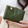 A hand holds a dark olive green minimalist card holder wallet with gold hardware against a light background.