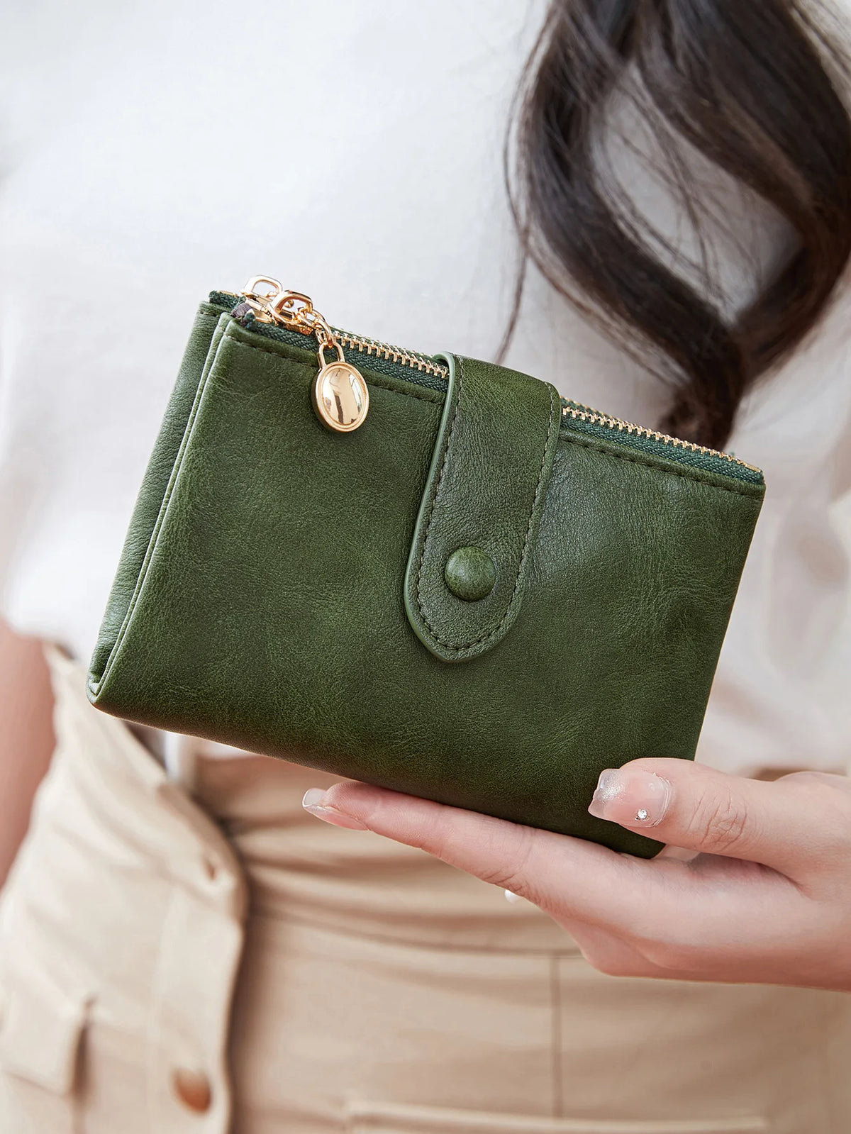 A hand holds a dark olive green minimalist card holder wallet with gold hardware against a light background.