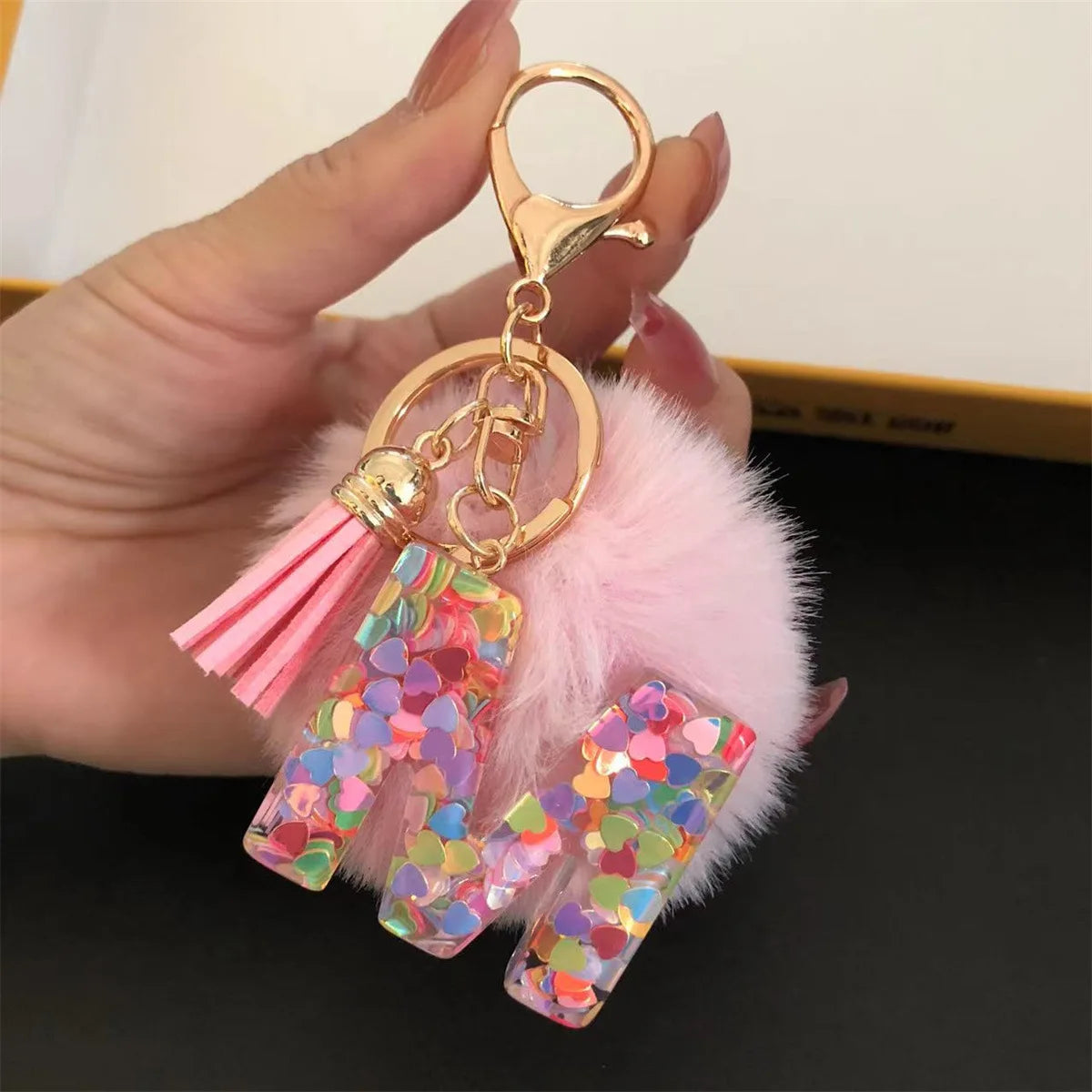 A gold-colored keychain featuring a pink fluffy pom-pom and a resin pendant shaped like the letter I.