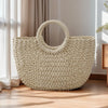 A large, light beige handwoven rattan beach bag resting on a wooden floor next to sheer curtains.
