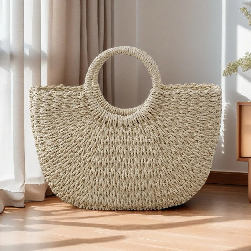 A large, light beige handwoven rattan beach bag resting on a wooden floor next to sheer curtains.