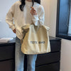 A woman wearing a white sweatshirt holds a beige canvas tote bag with blue lettering in front of a dark cabinet.