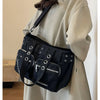 A black nylon shoulder bag with multiple straps and rivets is carried by a woman wearing a beige coat.