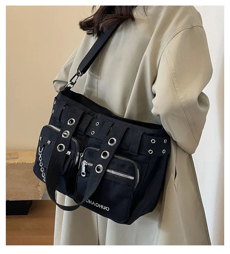 A black nylon shoulder bag with multiple straps and rivets is carried by a woman wearing a beige coat.