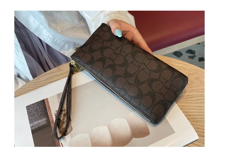 A dark brown monogrammed leather wallet with a gold zipper is shown resting on a light-colored surface.