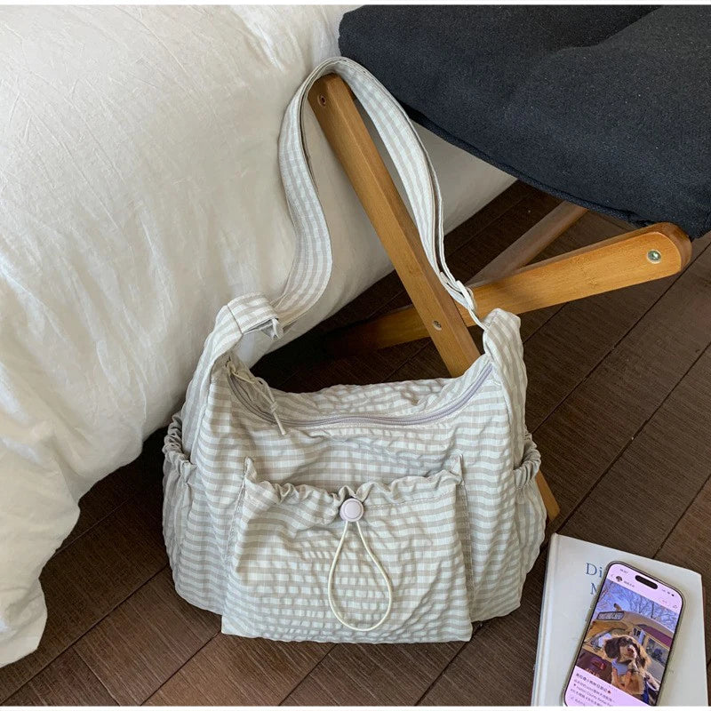 A light beige plaid nylon drawstring tote bag resting on a wooden surface next to a white pillow.