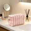 A pink and white striped waterproof travel cosmetic bag resting on a white countertop next to a mirror and brush.