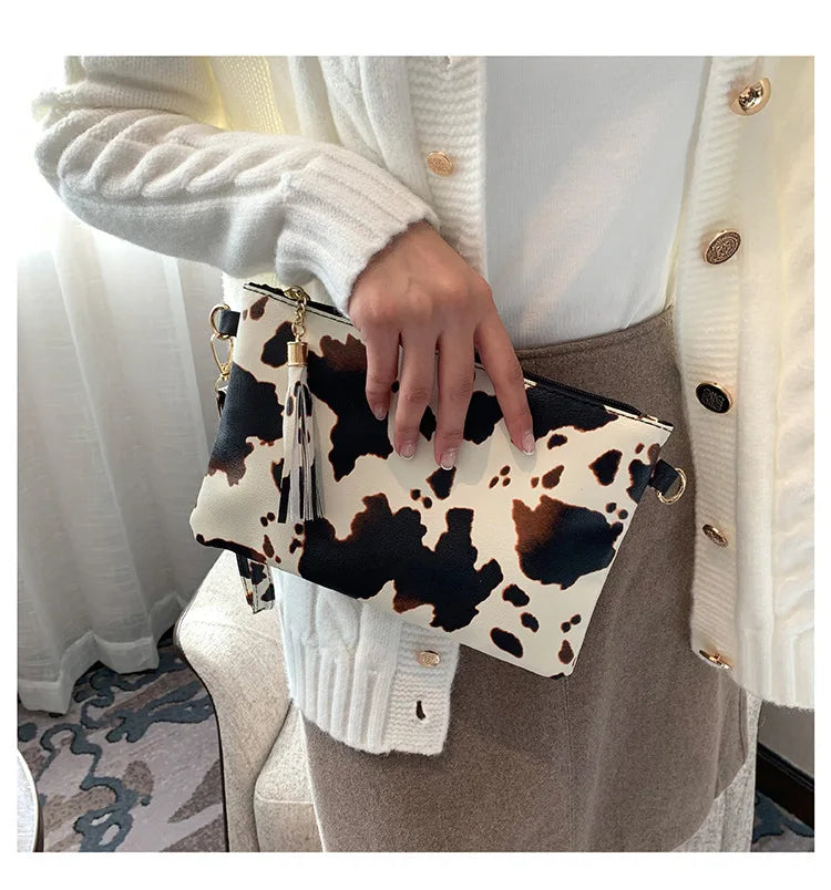 A hand holds a beige leopard print faux leather wrist bag against a white knitted sweater and tan skirt.