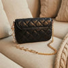 A black quilted flap bag with a gold chain extender is resting on a beige sofa cushion.