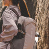A person wearing a light gray hoodie and dark hat carries a large beige hiking backpack against a textured rock face.