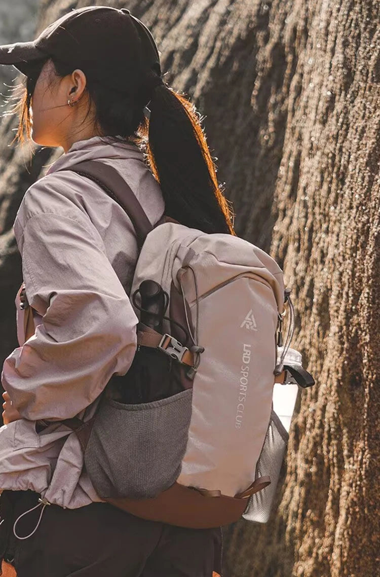 A person wearing a light gray hoodie and dark hat carries a large beige hiking backpack against a textured rock face.