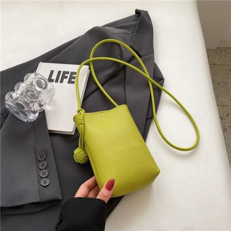 Bolso cruz de cuero verde lima con correa larga junto a un blazer gris y un accesorio transparente.