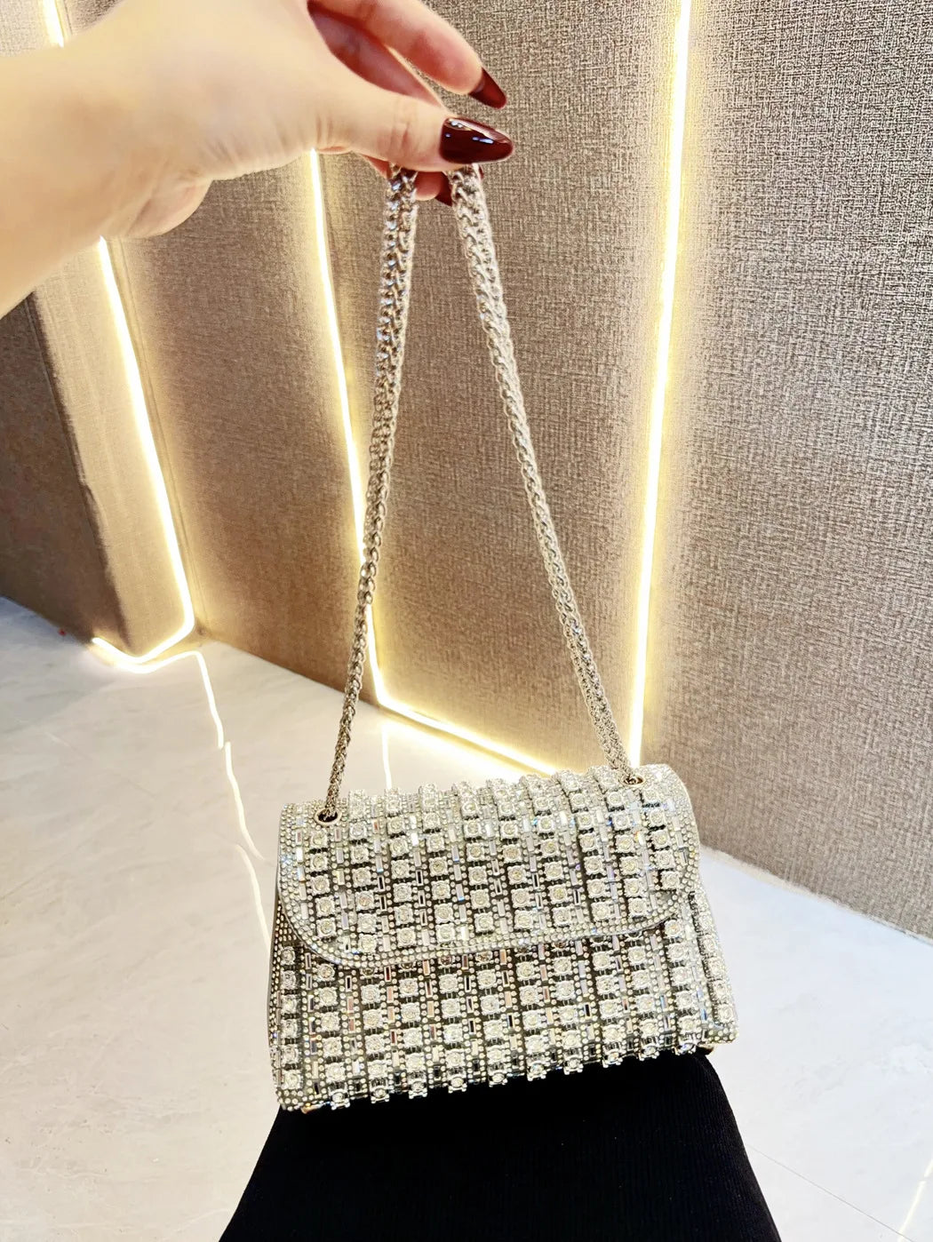 A hand holds an elegant rhinestone leather evening clutch bag against a textured light background.