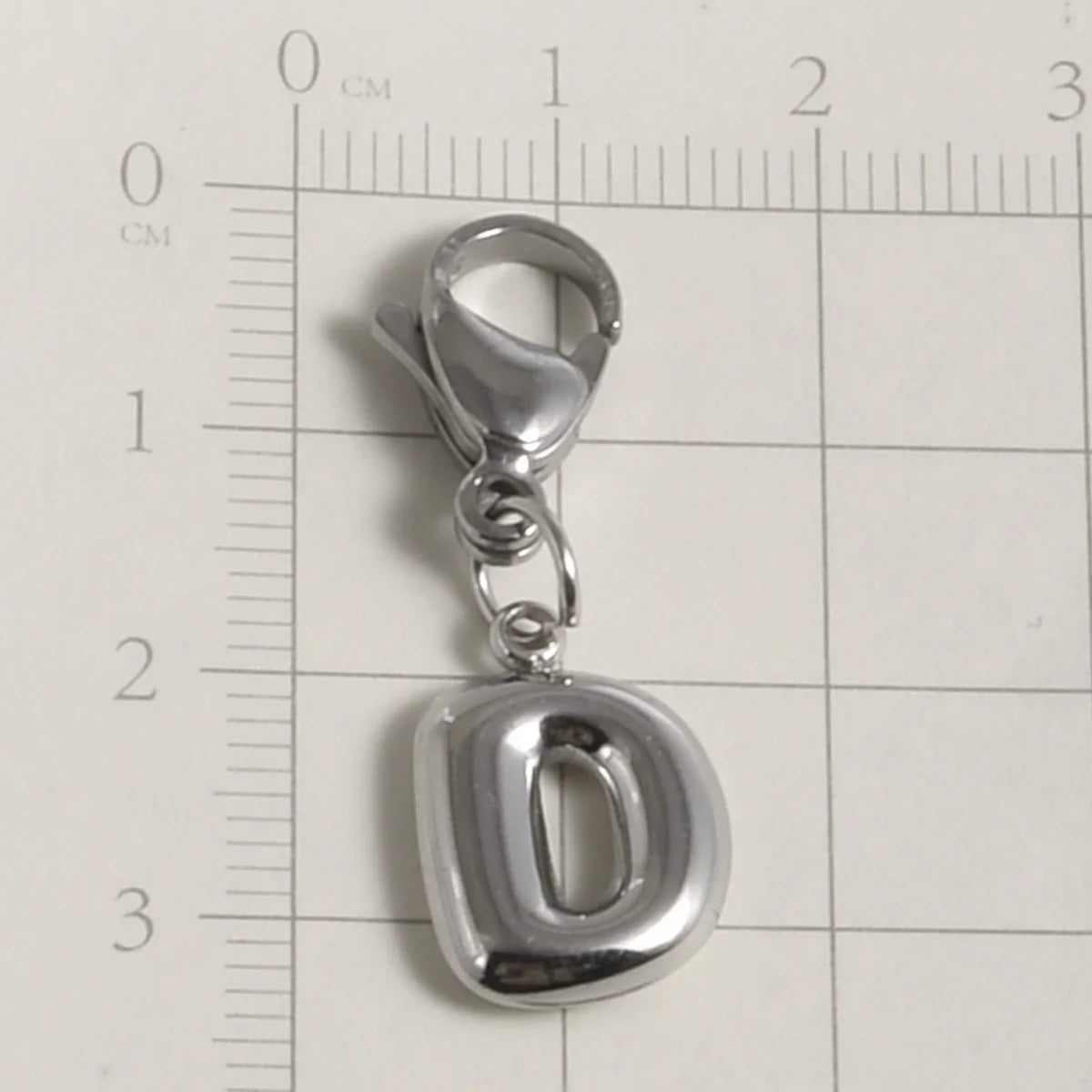 Silver stainless steel alphabet letter charm D with a lobster clasp shown on a ruler.