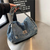 A denim tote bag with a chain strap is held by a hand against a dark background.