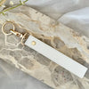 A white leather wristlet keychain with a gold-toned ring attached, resting on a marbled stone surface.