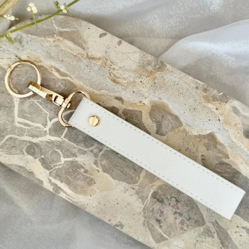 A white leather wristlet keychain with a gold-toned ring attached, resting on a marbled stone surface.