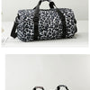 Maleta de viaje tipo duffel con estampado de leopardo en blanco y negro y correas negras, mostrada sobre una superficie blanc