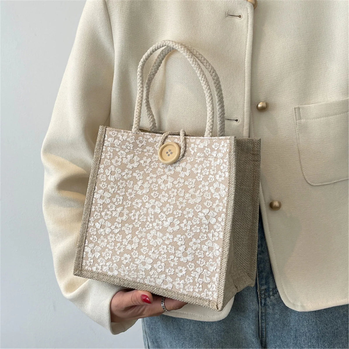 A beige linen tote bag with a floral pattern and secure closures is held by a person wearing a light jacket.
