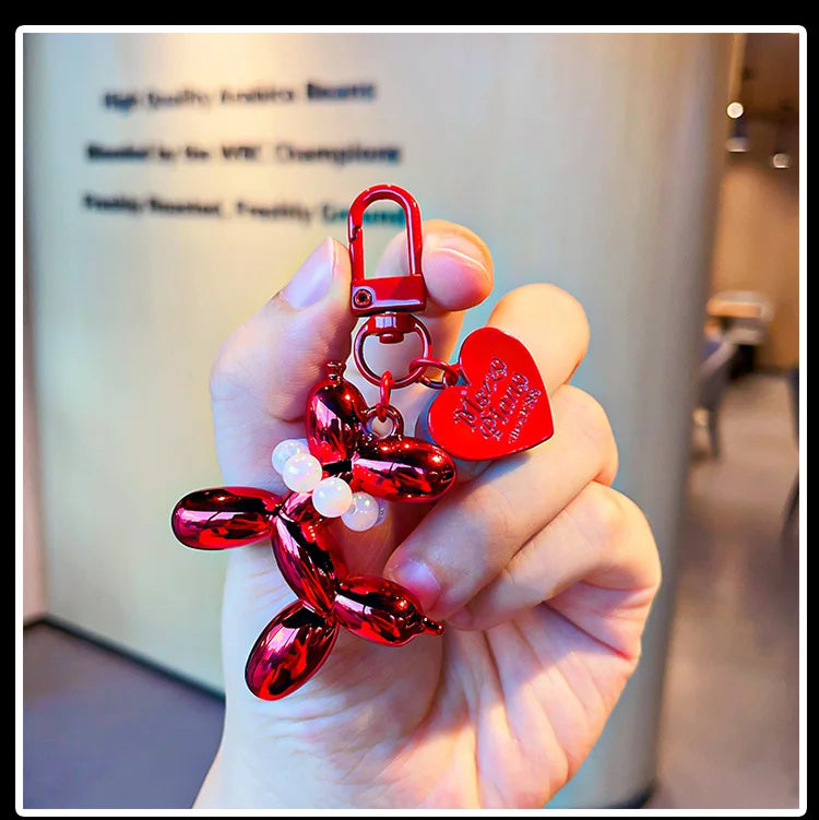 A hand holds a red balloon dog keychain charm with a heart pendant against a light background.