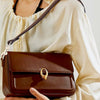 A brown leather handbag with a gold clasp is held by a person wearing a cream-colored blouse.