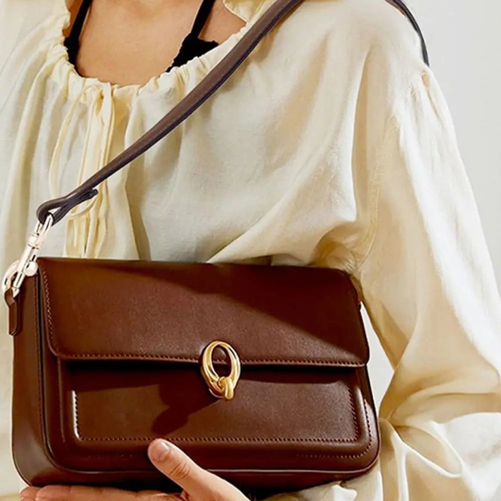 A brown leather handbag with a gold clasp is held by a person wearing a cream-colored blouse.