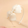 A close-up shot of a cream-colored bow hairband on a white elastic band against a light beige background.