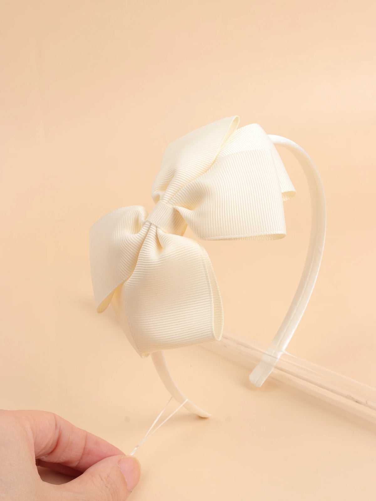 A close-up shot of a cream-colored bow hairband on a white elastic band against a light beige background.